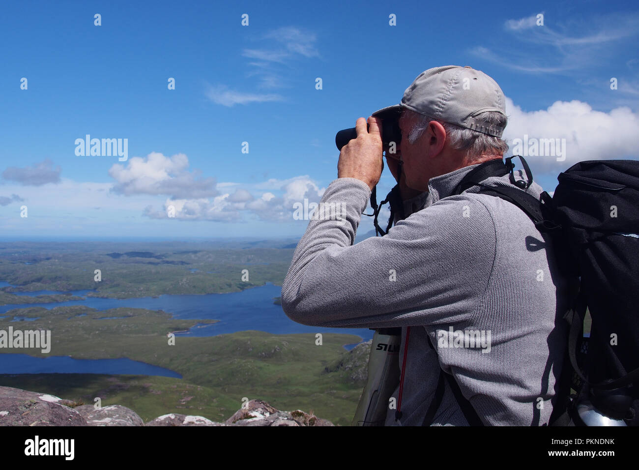 Homme d'une soixantaine de binoculars portant une casquette et sac à dos à partir de la Stac Pollaidh Inverpolly en forêt sur une journée ensoleillée Banque D'Images
