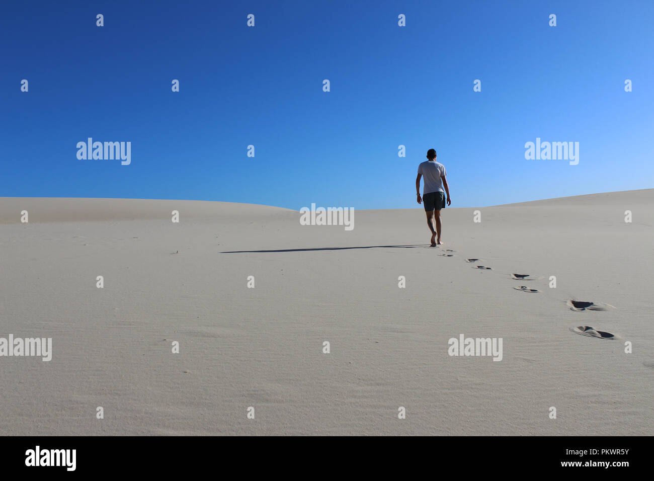 Homme sous le soleil brûlant dans un ciel bleu clair marchant sur une dune de sable blanc laissant ses empreintes dans le sable Banque D'Images