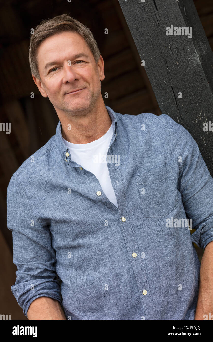 Portrait d'une belle réussite et heureux homme homme d'âge moyen portant une chemise bleue s'appuyant sur un poste par un garage ou grange Banque D'Images
