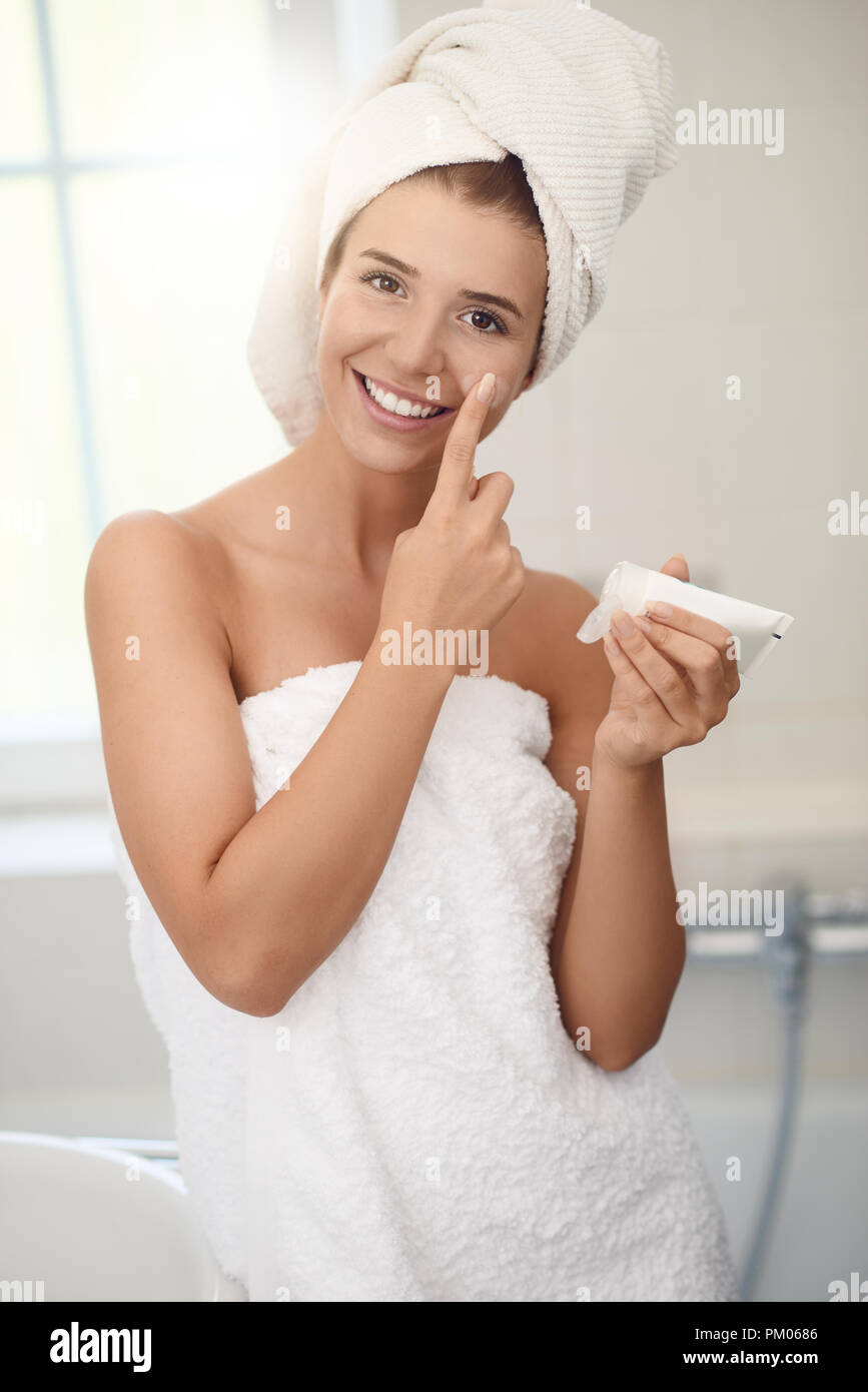 Jolie jeune femme en blanc serviettes dans une salle de bains donnant l'appareil photo un joli sourire alors qu'elle s'applique de la crème sur ses joues tandis que le bain en peau Banque D'Images