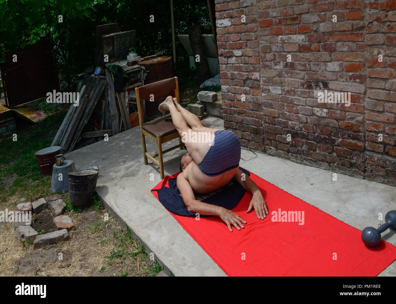 Homme chauve mature est en train de faire mentir branche dépasse l'exercice pour les abdominaux portant sur tissu rouge reste dans son arrière-cour près de mur de briques rouges en été Banque D'Images
