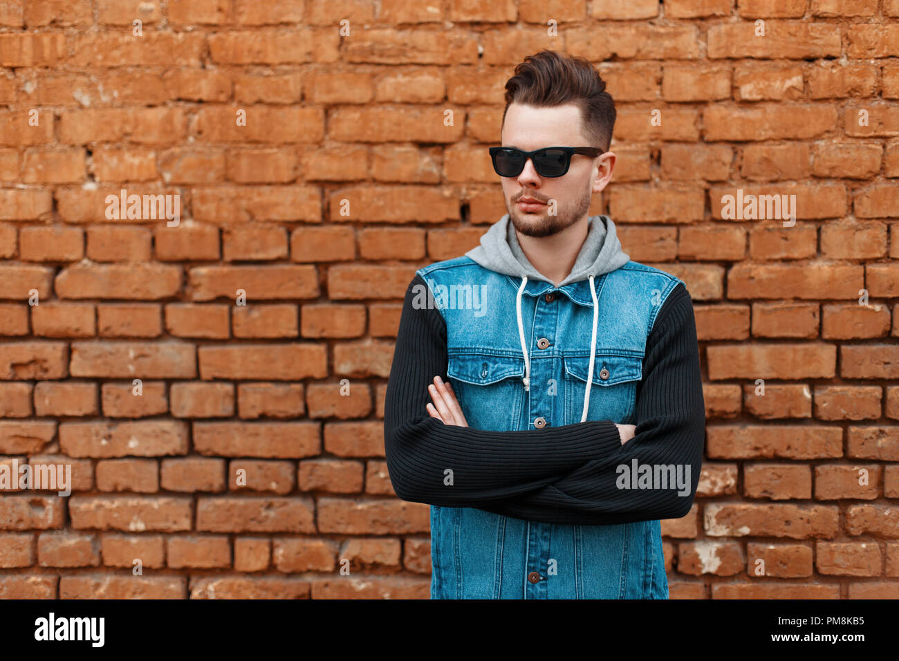 Bel homme à lunettes de soleil et un gilet en jean près d'un mur de briques Banque D'Images