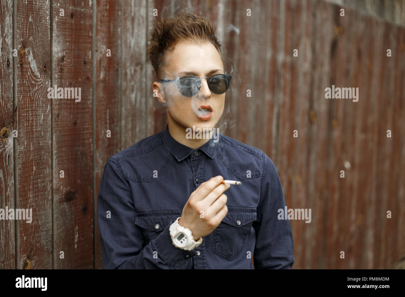 Jeune homme séduisant avec un souffle de cigarettes fumer à proximité d'un mur en bois vintage Banque D'Images