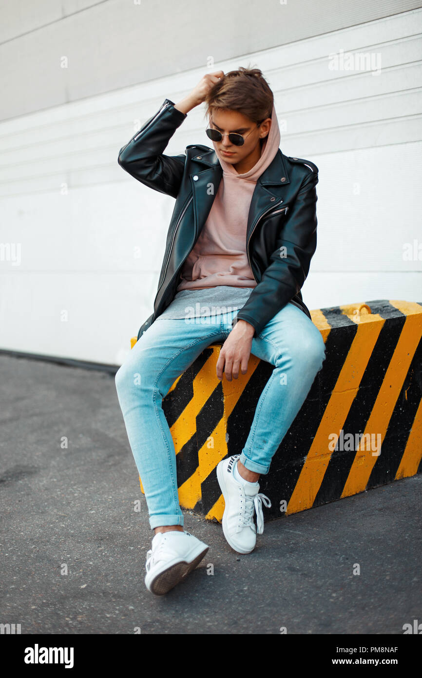 Beau jeune homme à la mode avec des lunettes de soleil dans une veste en  cuir noire, un jean bleu et blanc sneakers met sur un capot assis sur un  fond noir