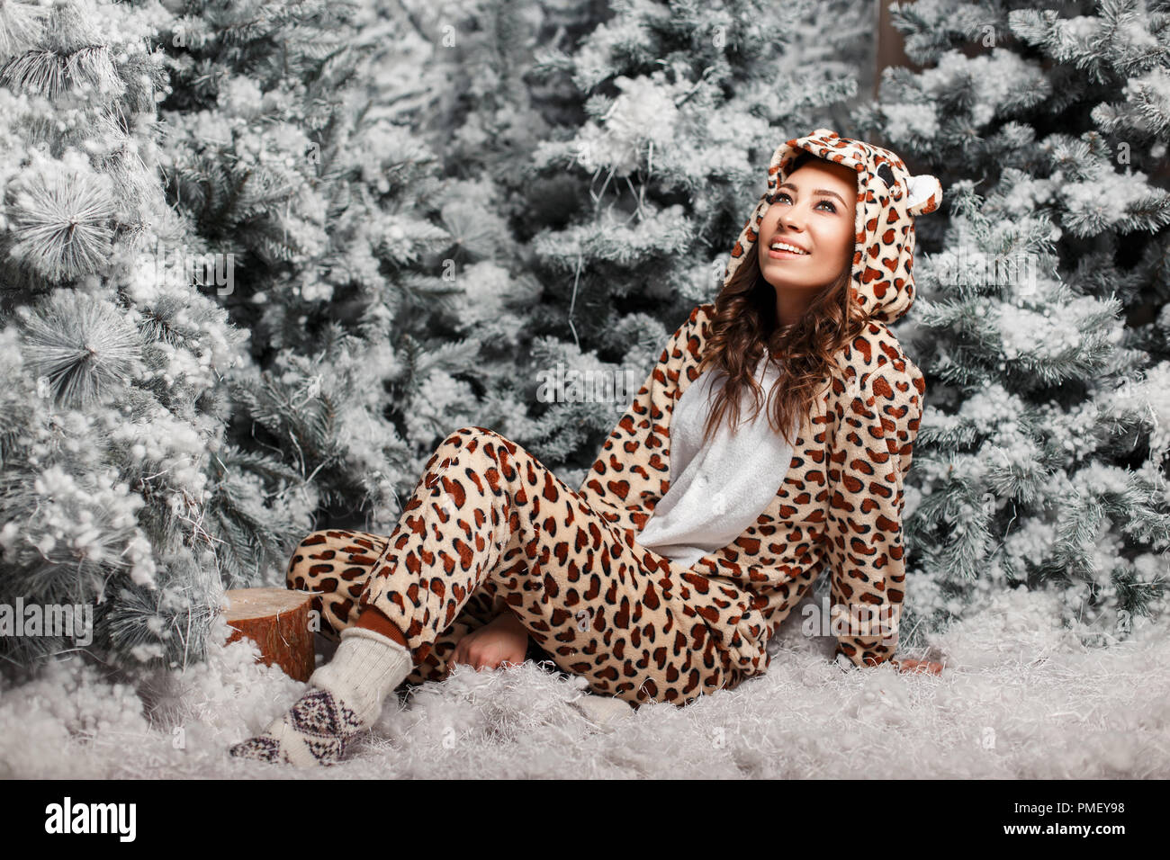 Jeune femme heureuse avec un sourire dans un drôle de pyjama ours se reposant près d'un arbre de Noël avec de la neige Banque D'Images
