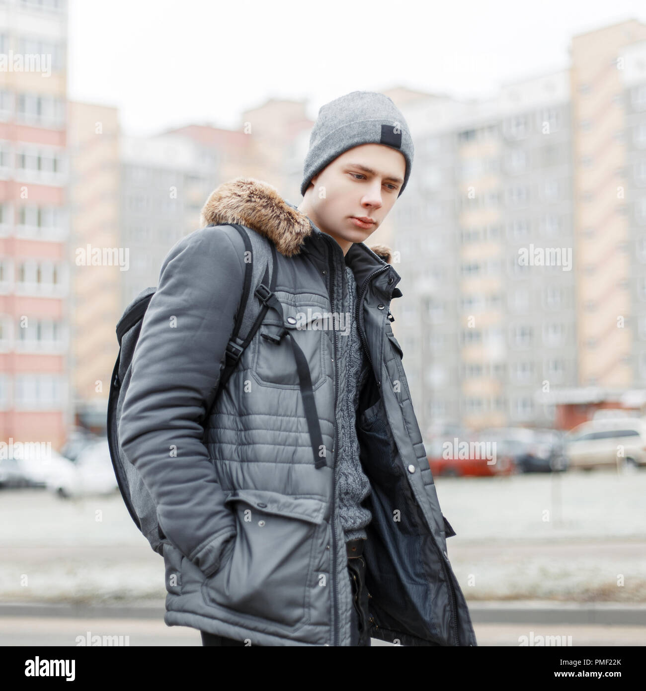 Jeune homme à la mode élégante dans une veste de mode d'hiver avec un  chapeau et un sac à dos voyages dans la ville dans le contexte de bâtiments  Photo Stock -