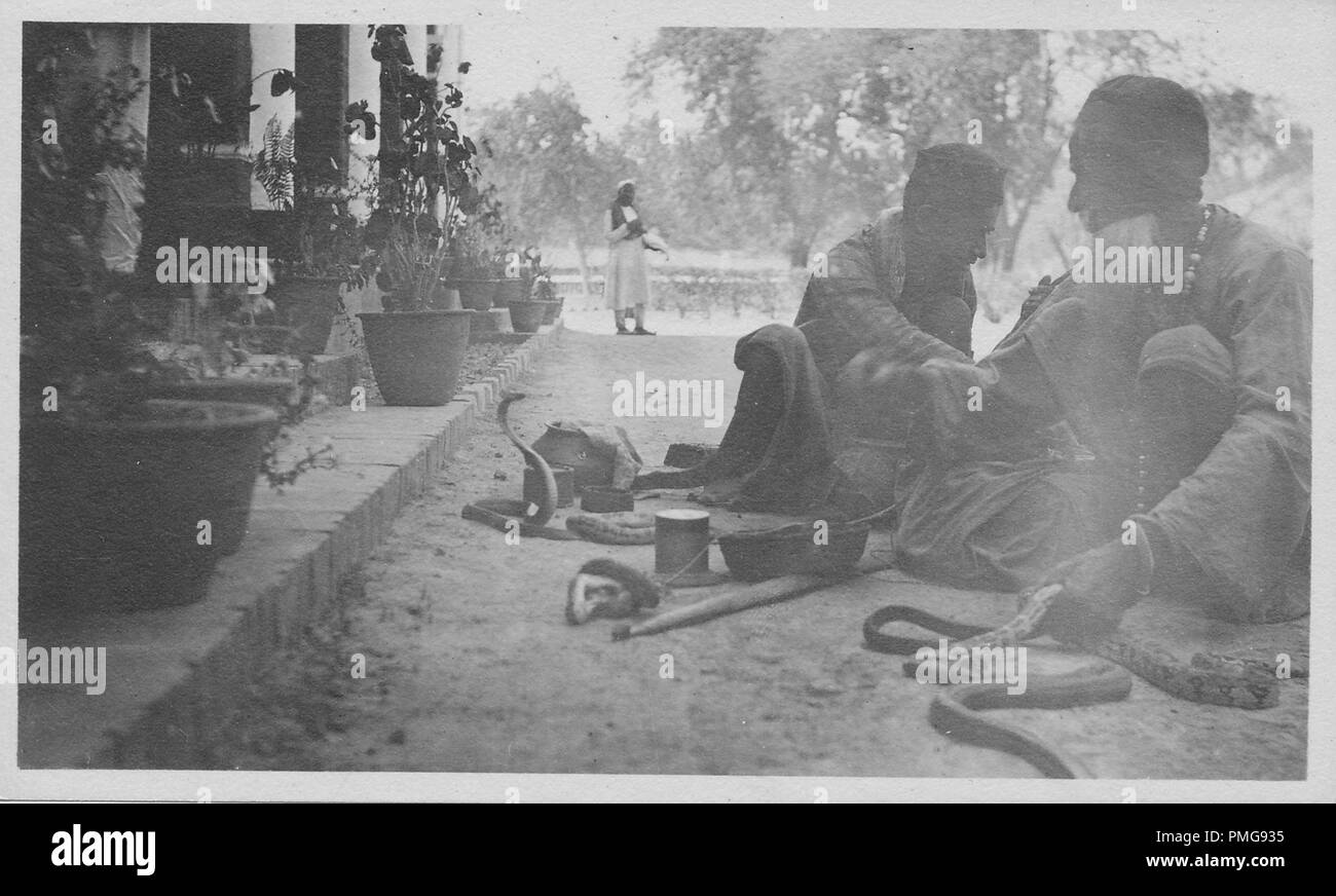 Photographie en noir et blanc sur papier cartonné, avec une image, tourné à partir d'un angle faible, de deux hommes, chacun portant une kurta, dhoti, et taqiya, assis sur le sol, avec plusieurs serpents et paniers, à côté d'une longue colonnade avec des plantes en pot, avec un homme portant un turban blanc kurta et debout dans l'arrière-plan, probablement perçus comme un souvenir touristique lors d'un voyage en Asie du Sud (Inde), 1910 probablement. () Banque D'Images