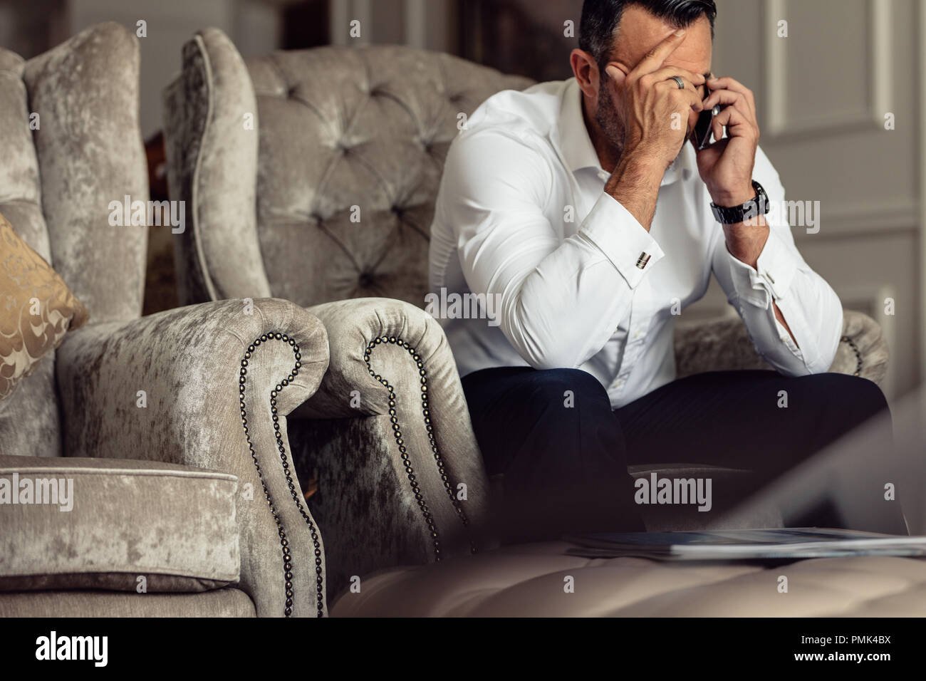 Portrait of a souligné mature businessman sitting in a hotel room et parlant au téléphone. Businessman sitting on couch with hand on head on cell p Banque D'Images