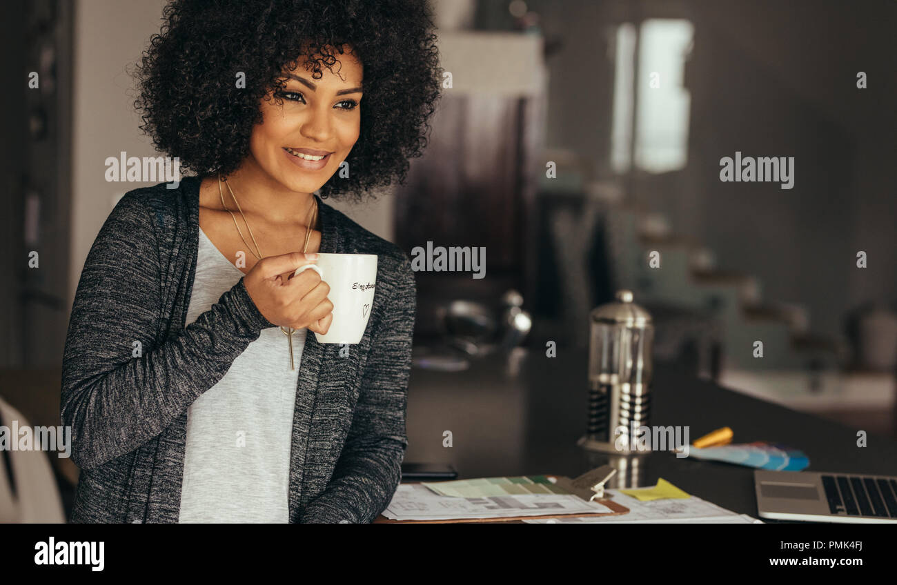 Happy woman taking pause café tout en travaillant au bureau à domicile. Femme africaine assise à son bureau avec une tasse de café à la voiture en souriant. Banque D'Images