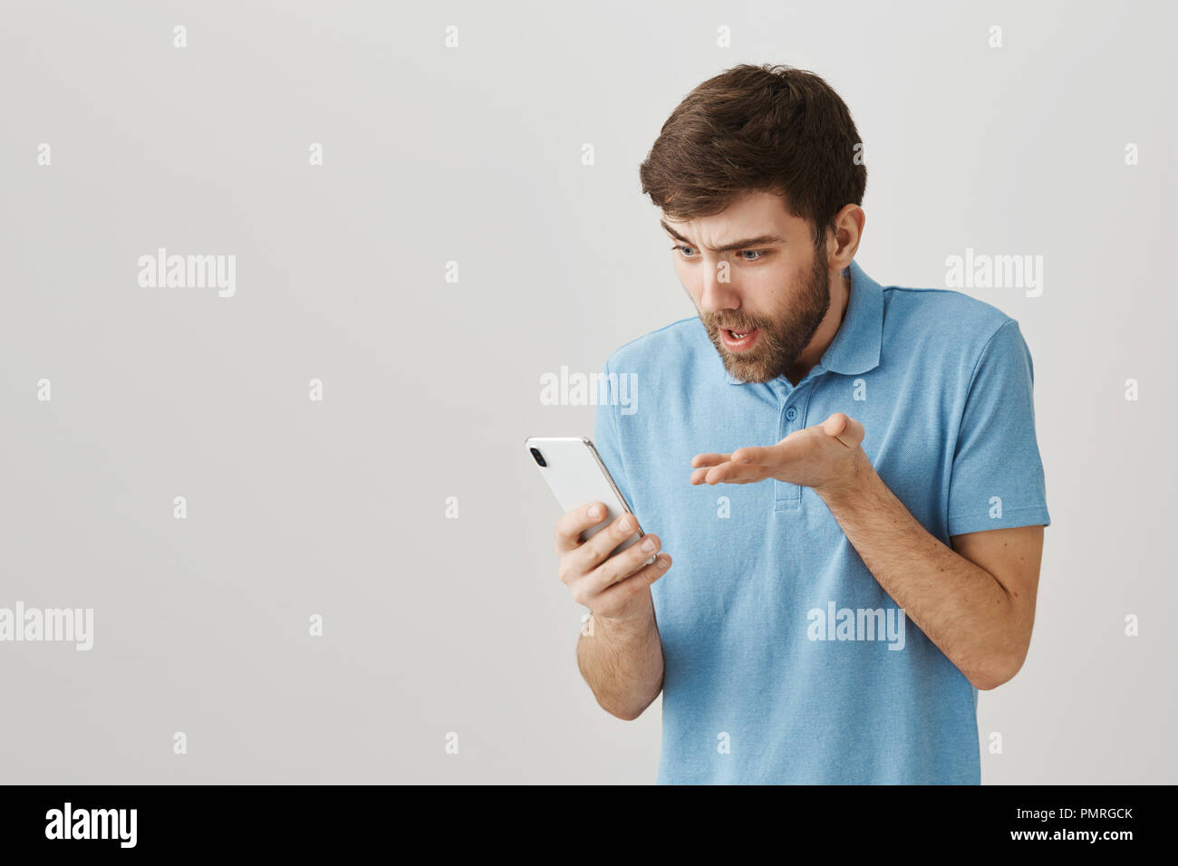 Qui vous a enseigné la grammaire. Studio shot of agacé et expressive portrait guy avec barbe holding smartphone tout en regardant l'écran et main tremblante, l'expression de la gêne et de la confusion Banque D'Images