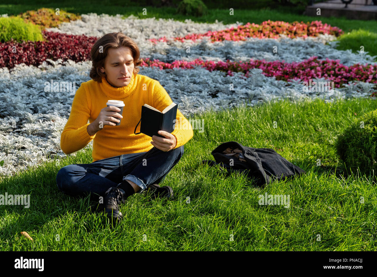 Le jeune homme boit du café et lit le livre dans le parc, il sourit, a un reste, est vêtu d'un chandail jaune, une veste imperméable ou noir, jeans Banque D'Images