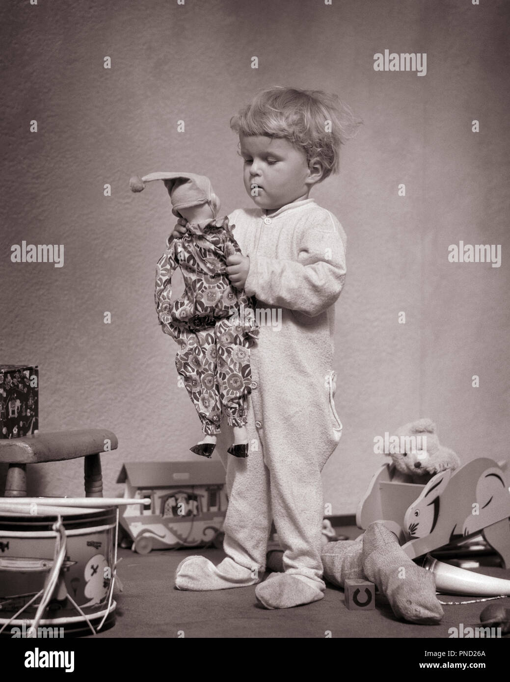 1930 JEUNE BLOND Bébé garçon debout portant des chaussettes de laine PUTOIS  DR. DENTON SLEEPWARE tenant une poupée favorite JESTER AU MILIEU D'AUTRES  JOUETS - j4508 HAR001 COPIE HARS pleine longueur de