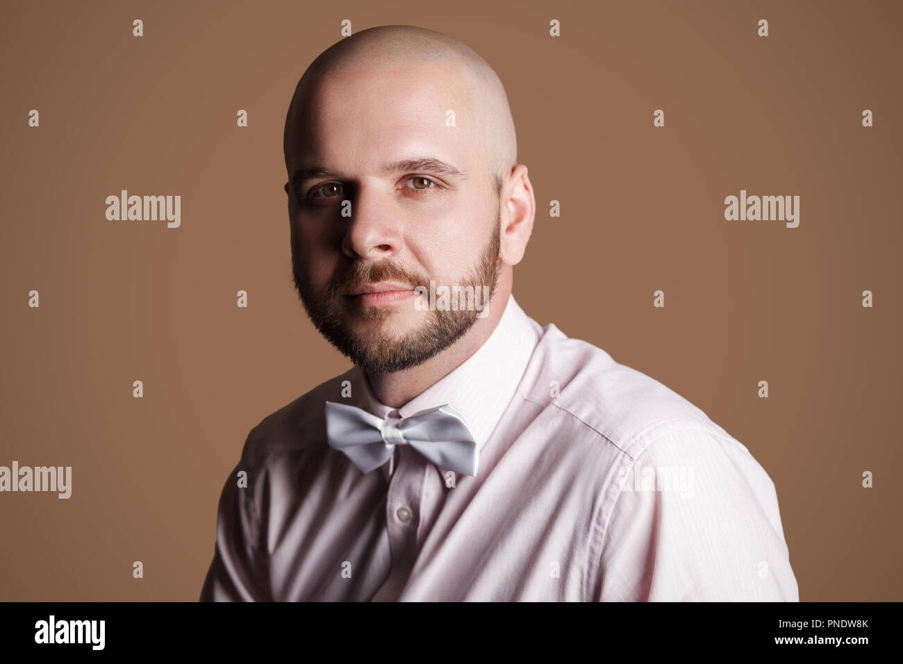 Closeup portrait of handsome man chauve barbu en chemise rose et arc blanc, assis sur une chaise et looking at camera with convaincu convaincu face. Banque D'Images