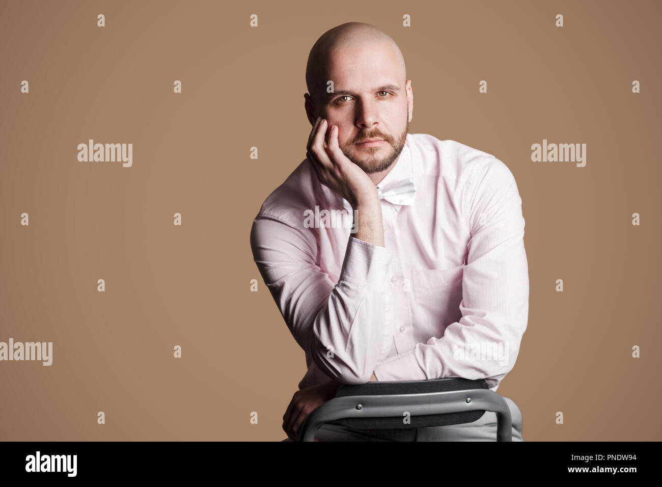 Portrait de barbu beau sérieux homme chauve en chemise rose clair et arc blanc, assis sur une chaise et looking at camera with hand on chin. Piscine stud Banque D'Images