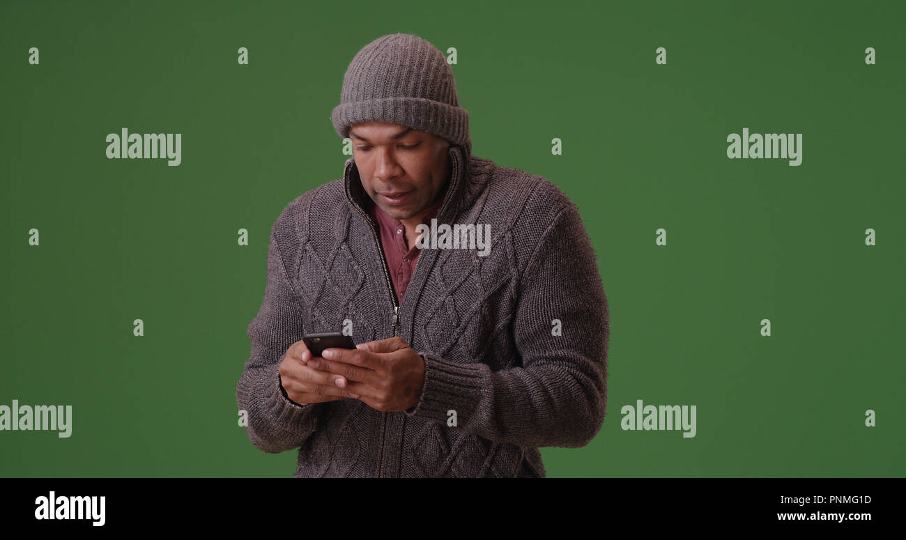 African American man in beanie et sweater texting on téléphone sur un écran vert Banque D'Images