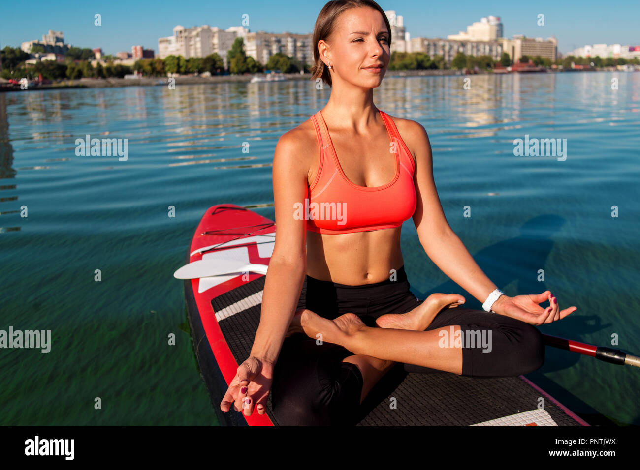Stand Up Paddle board yoga effectué par belle fille sur la ville, l'arrière-plan lumineux sur l'eau formation de yoga Banque D'Images
