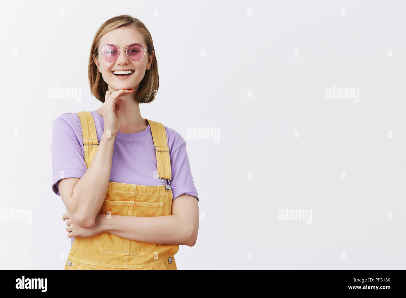 Mignon femelle magazine editor dans le quartier branché de lunettes de soleil et une salopette jaune, touchant le menton et holding hand croisées sur la poitrine, debout dans intrigué et curieux pose, avoir été impressionné par le guitariste de rue Banque D'Images