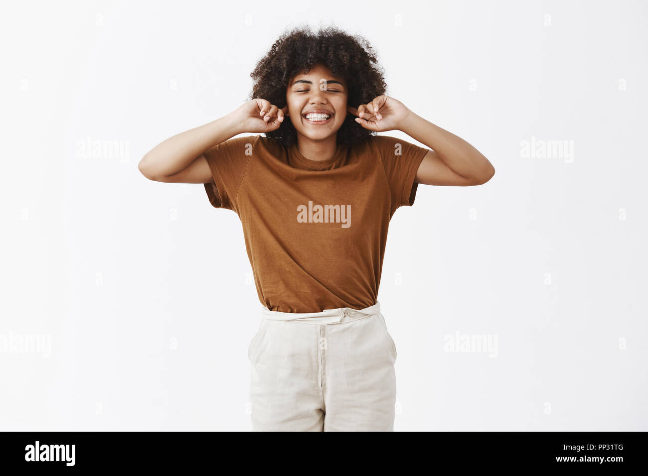 Insouciants et heureux à la peau sombre attrayant étudiante avec les cheveux bouclés en costume élégant fermer les yeux et sourire insouciant avec expression indifférente couvrant les oreilles avec l'index Banque D'Images