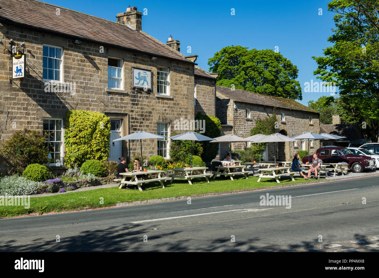 Les clients bénéficient de boissons en plein air au soleil sous les parasols tables à l'extérieur de la pittoresque Blue Lion Inn - East Witton, North Yorkshire, Angleterre, Royaume-Uni. Banque D'Images