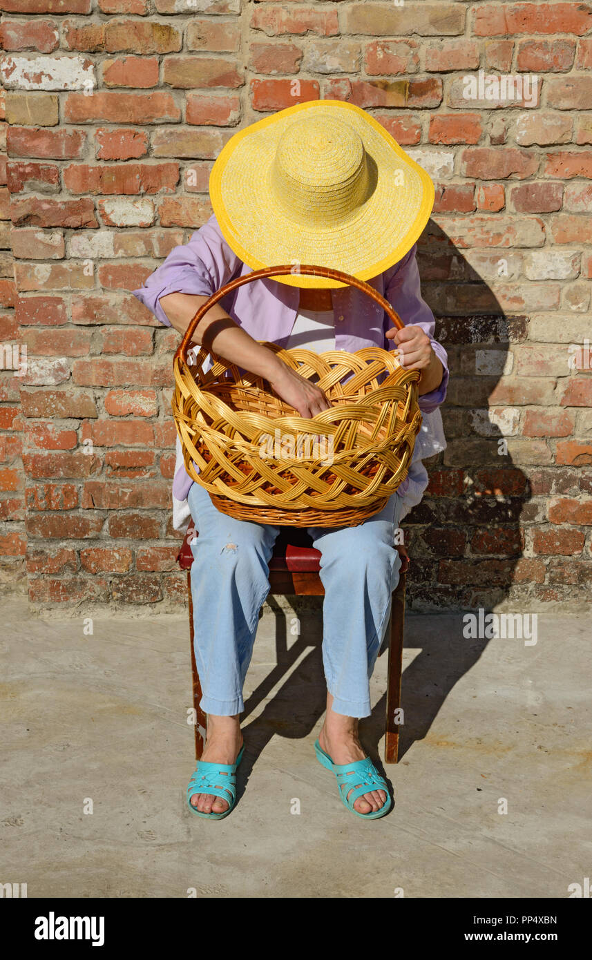 Close-up portrait of mature woman in chapeau à large qui est assis sur la chaise baissant la tête plus grand panier en osier sur ses genoux en Banque D'Images