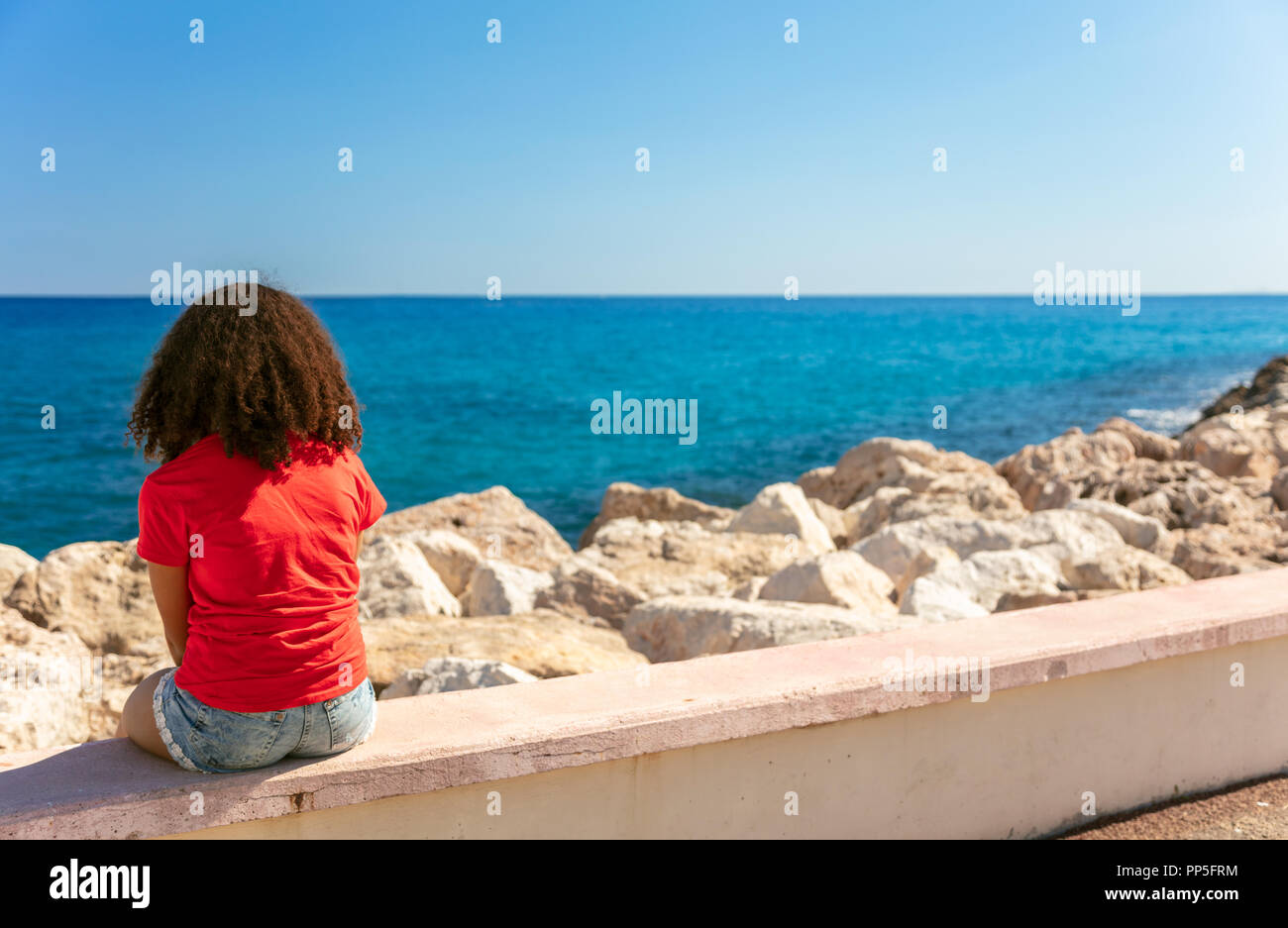 Mixed Race African American girl adolescent femme jeune femme portant un t-shirt et short en jean assis sur un mur donnant sur une mer bleue Banque D'Images