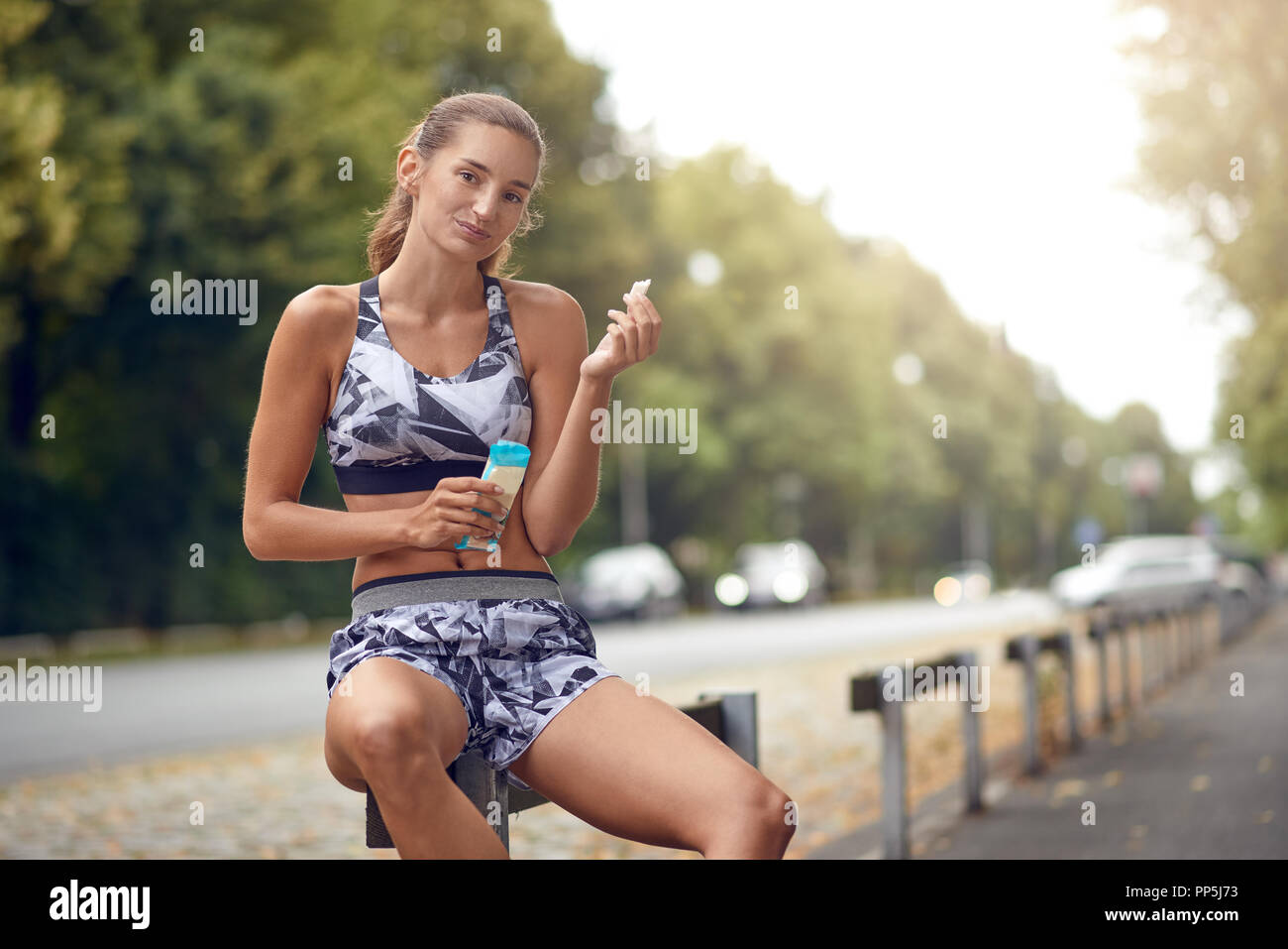 Belle jeune femme élancée sportive assis sur un rail route de manger un morceau d'une protéine bar alors qu'elle prend une pause dans son quotidien jog Banque D'Images