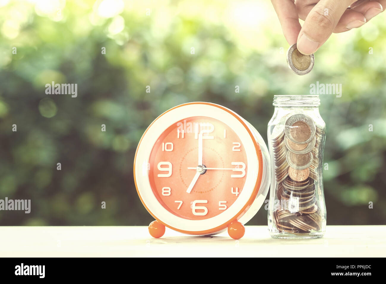 L'argent d'épargne et investissement de temps concept : un homme part mettre de monnaie dans un bocal en verre avec de l'horloge sur une table avec la nature verte comme arrière-plan. Concept Banque D'Images