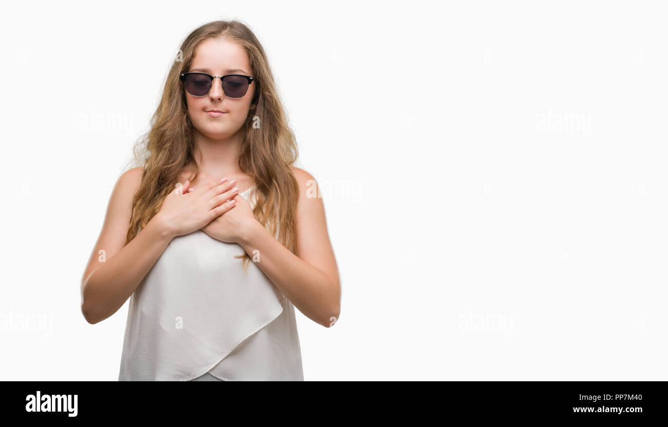 Young blonde woman wearing sunglasses smiling avec les mains sur la poitrine avec les yeux fermés et reconnaissant geste sur la face. Concept de santé. Banque D'Images