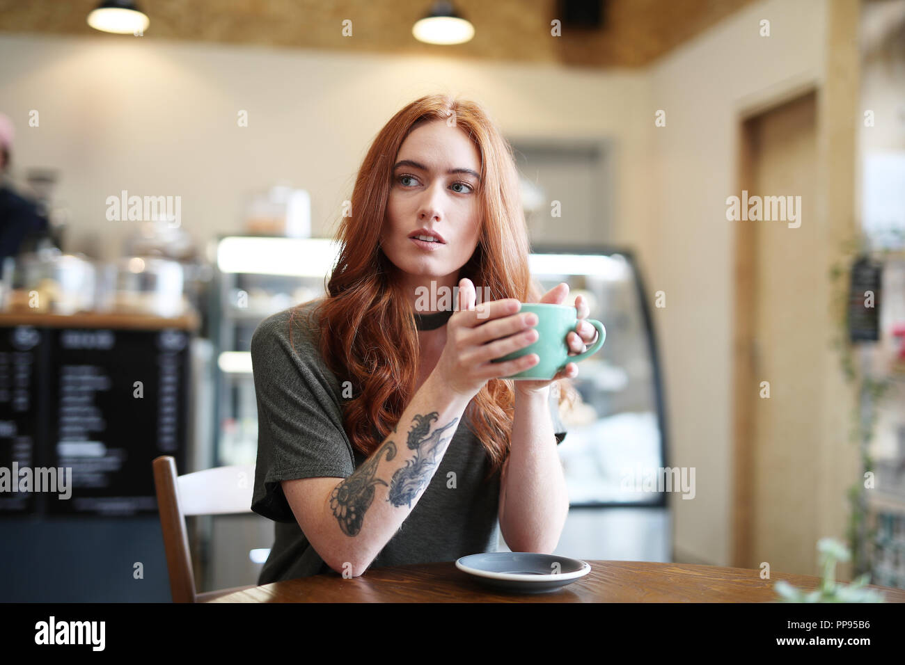 Une femme avec un tatouage bras est assis à une table en bois tenant une tasse de Cappuccino dans un bol vert Banque D'Images