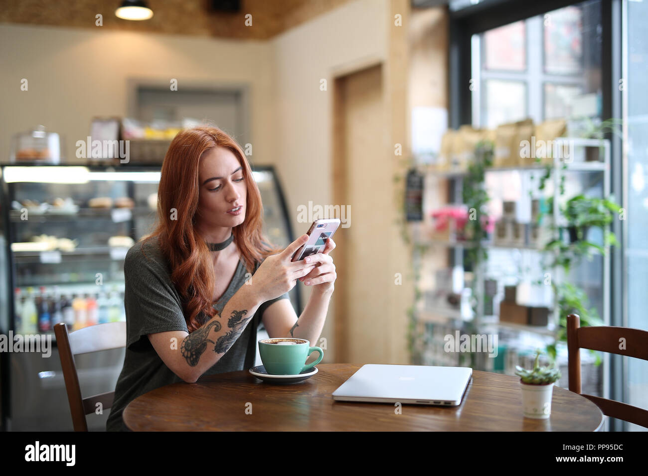 Red Head girl avec bras tatouage, assise seule dans un café en regardant son téléphone portable, avec un ordinateur portable assis sur la table en bois, Liverpool, Royaume-Uni Banque D'Images