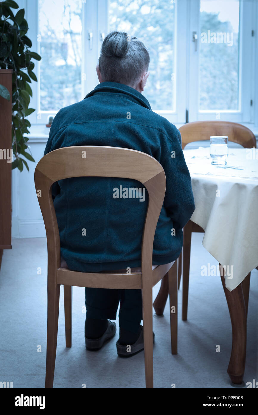 Vieille veuve assise seule à sa table du salon avec un verre d'eau en regardant par la fenêtre, l'effet de filtre bleu, concept de la dépression des personnes âgées Banque D'Images