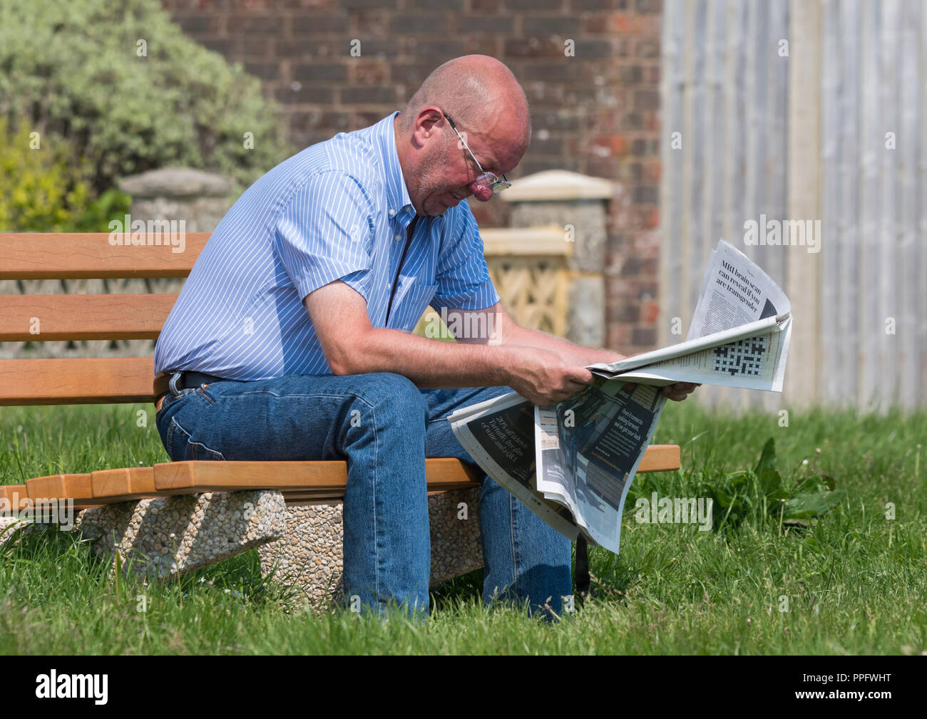 Smart habillé décontracté caucasian middle aged homme assis sur un banc de parc en bois de lire le journal dans le Royaume-Uni. Banque D'Images