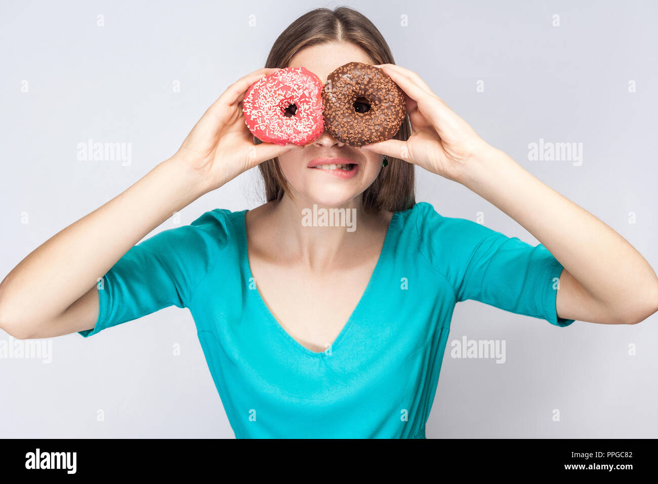 Jeune fille fou une morsure des lèvres en blouse bleue, tenant debout, couvrant les yeux avec des beignets au chocolat et rose,recherche par donuts comme la fabrication du verre Banque D'Images