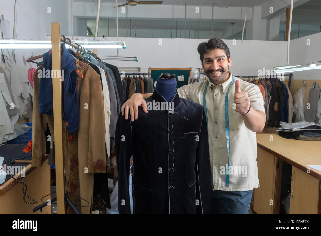 Adapter debout à côté d'un mannequin portant un manteau semi-cousues dans son atelier et showing Thumbs up sign. Banque D'Images Adapter debout à côté d'un mannequin portant un manteau semi-cousues dans son atelier et showing Thumbs up sign. Banque D'Images