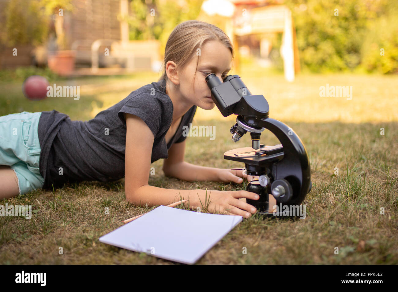 Une fillette de 9 ans se trouve sur l'herbe dans le jardin et regarde dans l'oculaire d'un microscope. En face d'elle est une feuille de papier. Banque D'Images