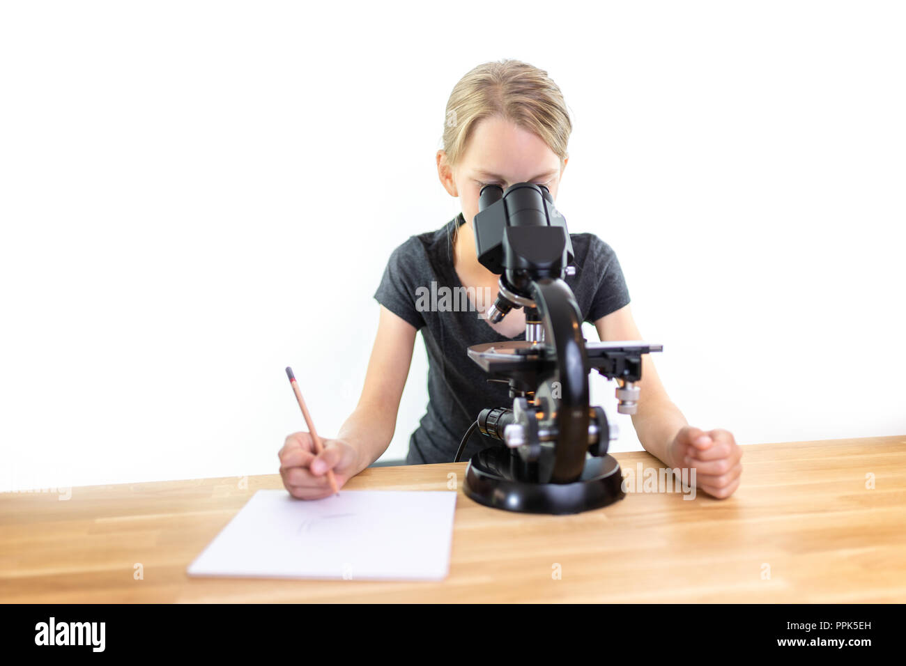 Une petite fille de 9 ans cherche dans un oculaire d'un microscope et note ses observations sur une feuille de papier. Contre isolé sur fond blanc Banque D'Images