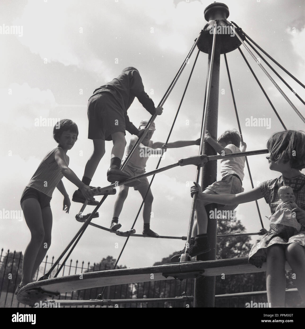 Années 1960, historiques, les jeunes enfants à l'extérieur dans un terrain accroché sur les traverses dans un passionnant metal swing ou merry-go-round ride, connu sous le nom de "Chapeau de sorcière", England, UK. Plus tard dans la décennie, ils ont été retirés de terrains en raison de préoccupations en matière de santé et de sécurité sur un enfant tomber entre eux. Banque D'Images