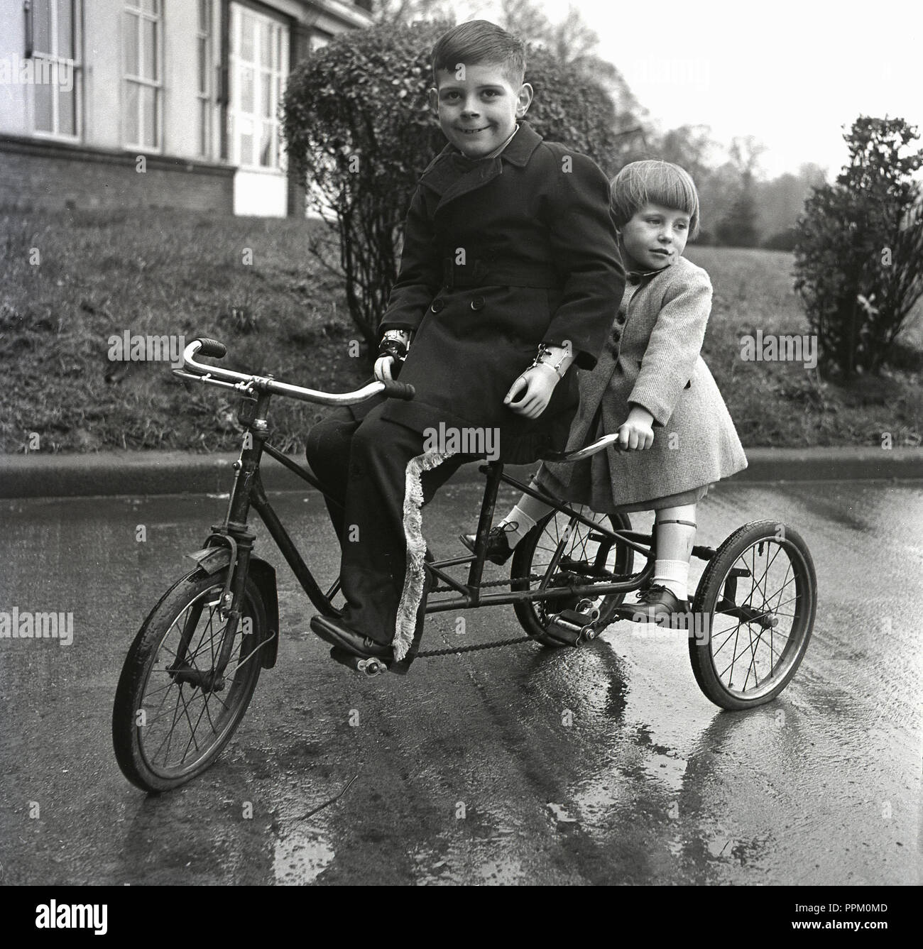 1950s, historique, deux jeunes enfants handicapés physiquement reçoivent de l'air frais lorsqu'ils s'assoient ensemble sur un tricycle en tandem à l'extérieur de leur hôpital, en Angleterre, au Royaume-Uni. Banque D'Images
