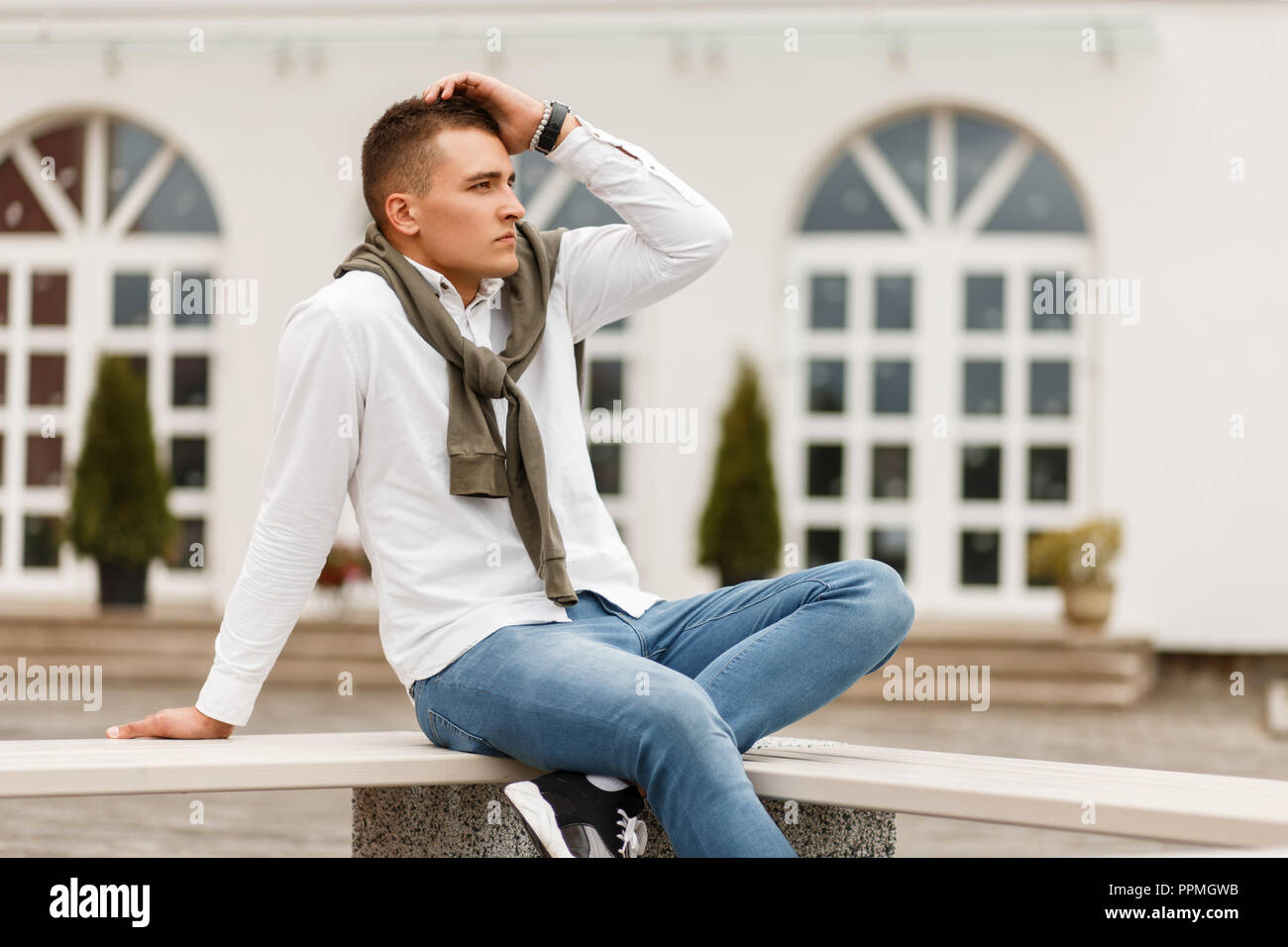 Beau succès à la mode homme en chemise blanche avec un chandail et un jean  assis sur un banc près de l'hôtel Photo Stock - Alamy