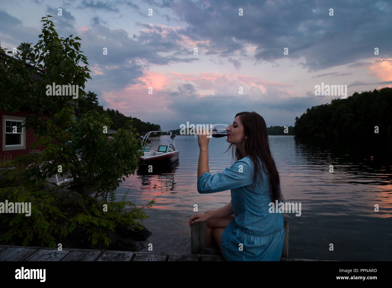 Jeune femme brune assise sur un quai au bord du lac au coucher du soleil un verre de vin rouge. Banque D'Images