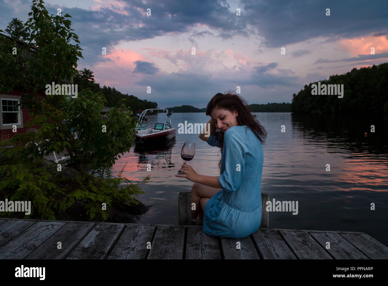 Jeune femme brune assise sur un quai au bord du lac au coucher du soleil un verre de vin rouge. Banque D'Images