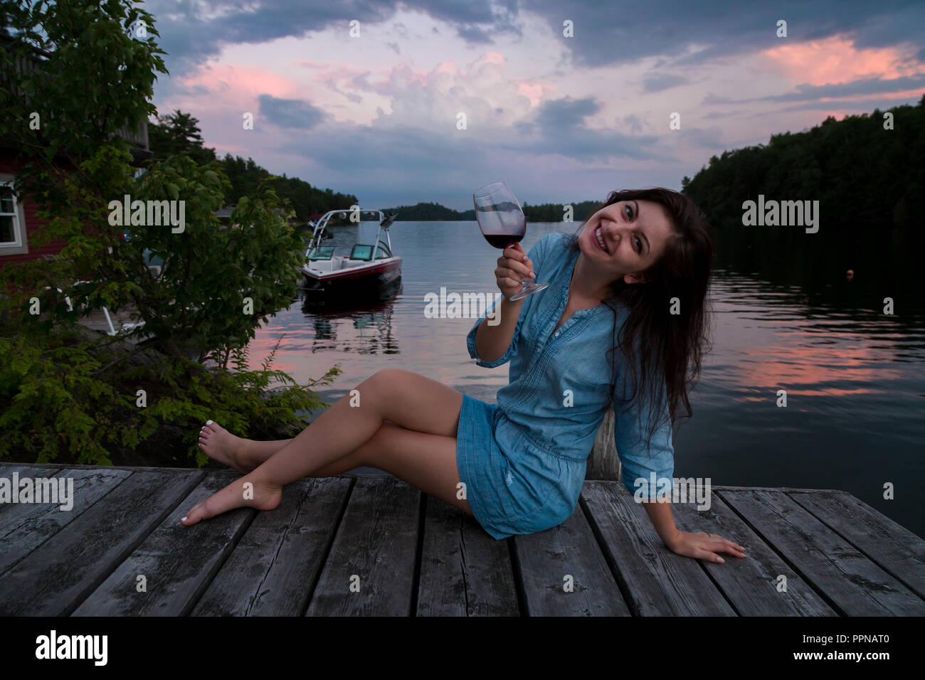 Jeune femme brune assise sur un quai au bord du lac au coucher du soleil un verre de vin rouge. Banque D'Images