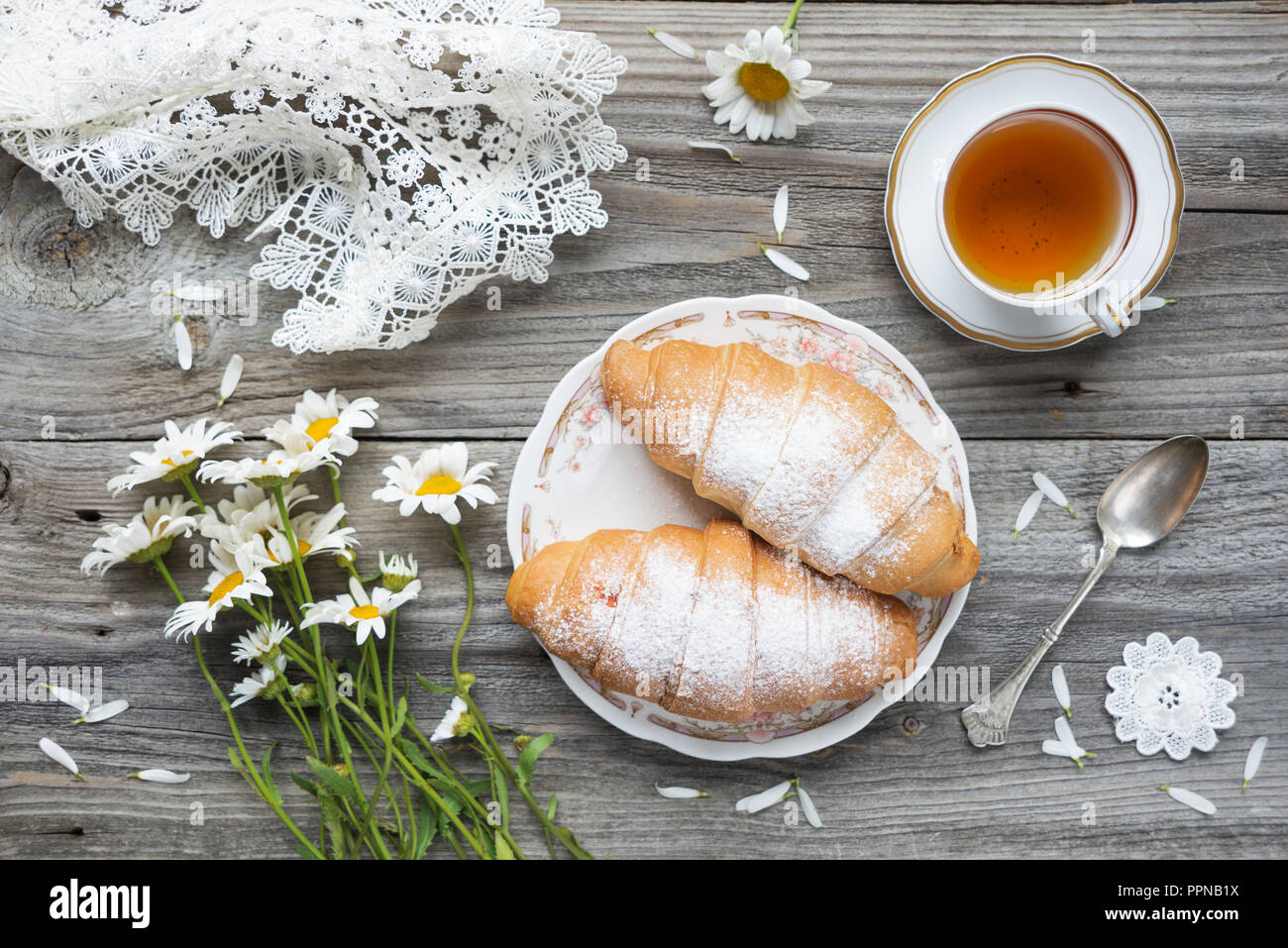 Belle composition : deux croissants, bouquet de fleurs Daisy, tasse de thé et blanc lacy nappes sont sur un vieux fond de bois Banque D'Images