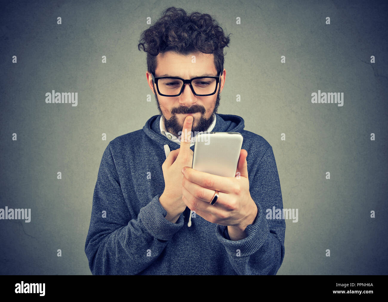 L'homme intrigué penser à ce que de répondre aux messages texte sur téléphone cellulaire isolé sur fond de mur gris. Le langage du corps de réaction expression visage Banque D'Images