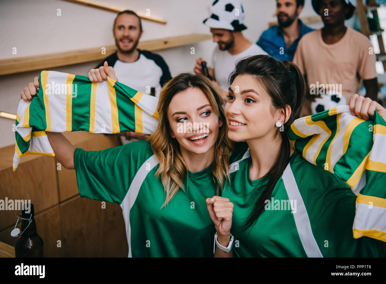 Les femmes en vert fan t-shirts holding foulard et leurs amis masculins debout derrière pendant veille de match de foot au bar Banque D'Images