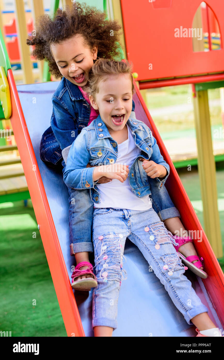 Deux excités hurler les petits enfants multiethnique glisser vers le bas à partir de la colline de jeux pour enfants Banque D'Images