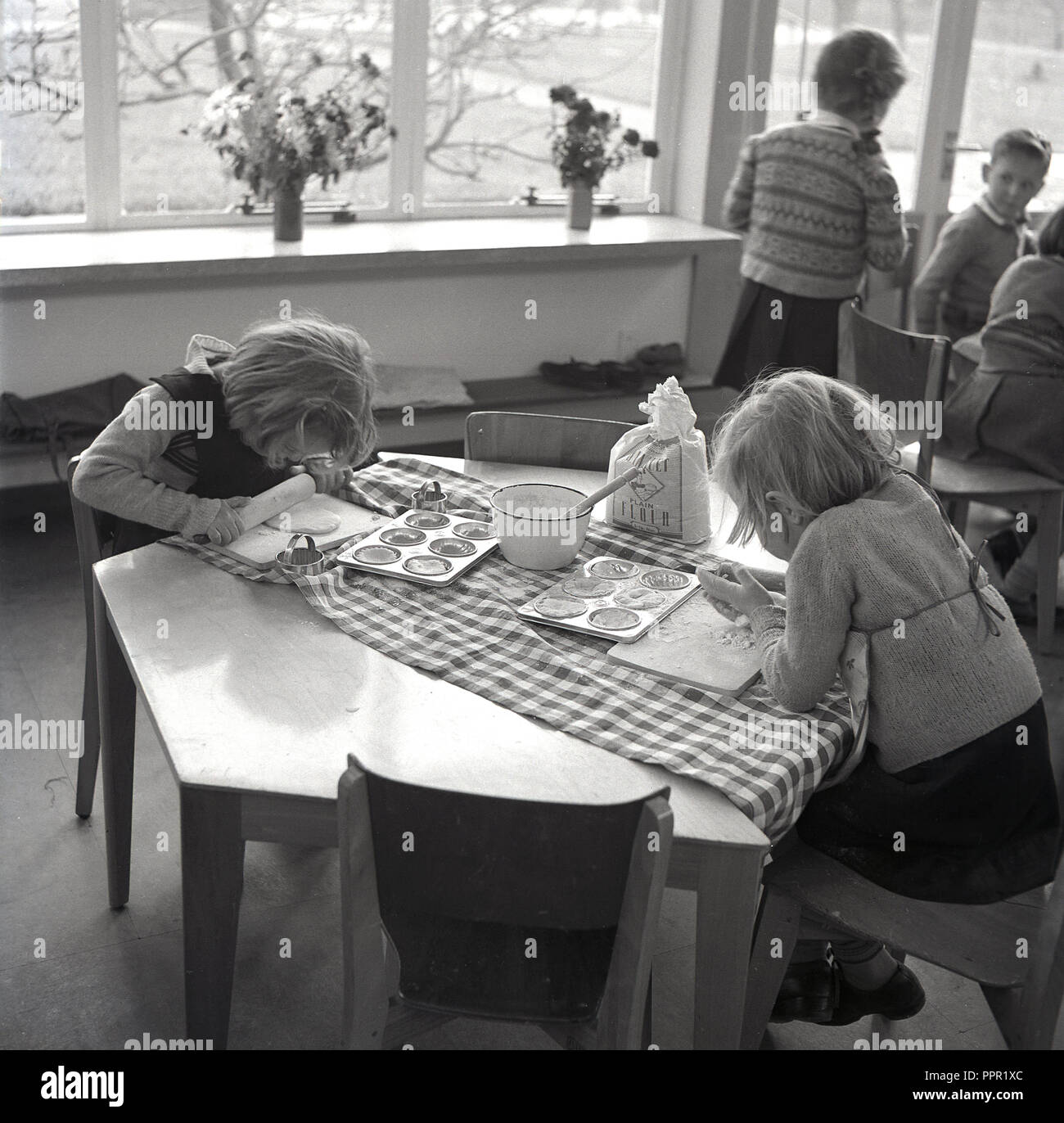 Années 1960, historiques, une école de cuisine, deux jeunes écolières assis à une table hexagonale avec un sac de farine et un bol d'eau en pâte. Un des girl's est à l'aide d'un matériel roulant en bois-pin pour aplatir la pâte, England, UK. Banque D'Images