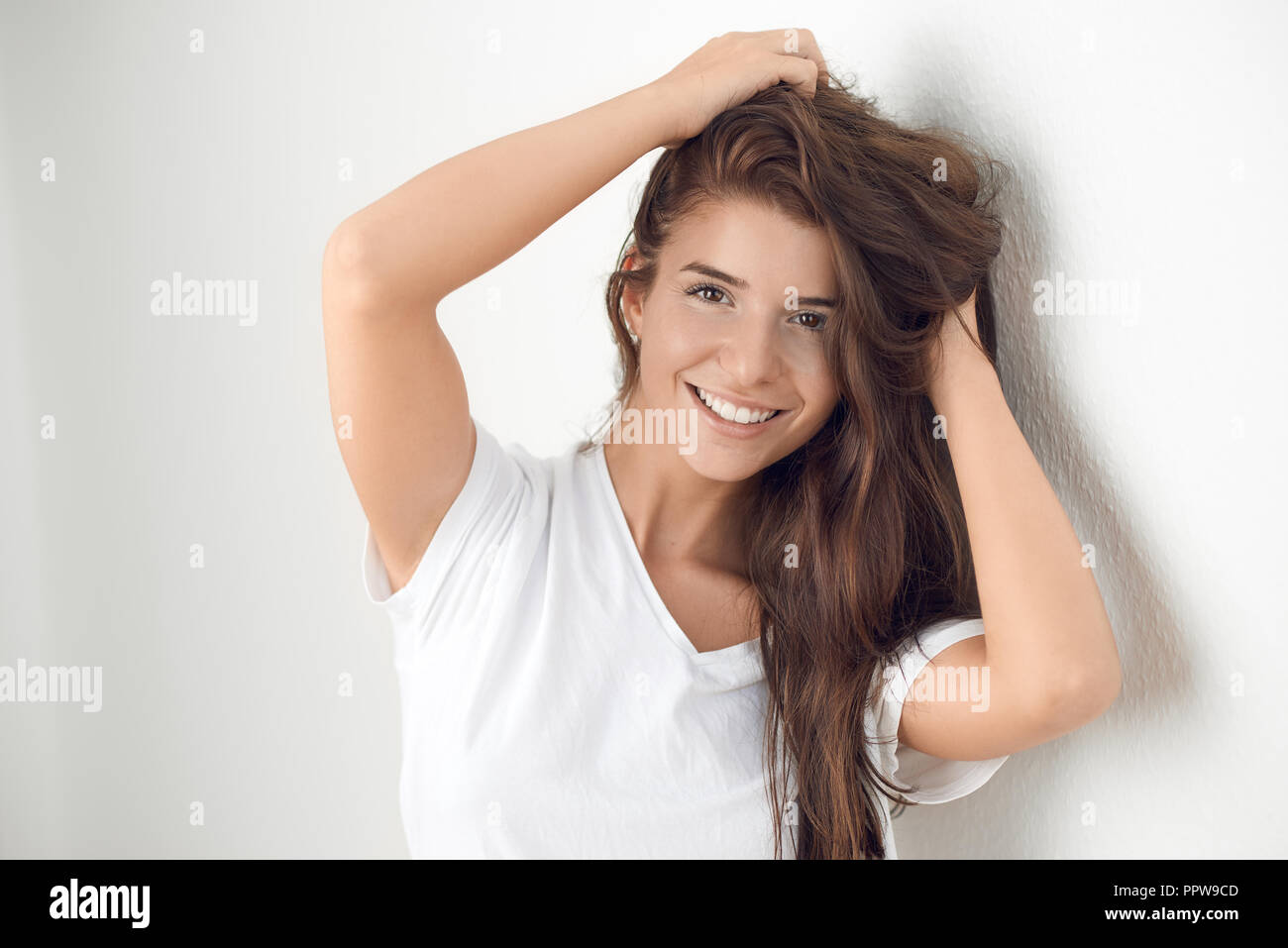 Young smiling blond woman holding ludique ses cheveux tout en se penchant contre mur blanc Banque D'Images