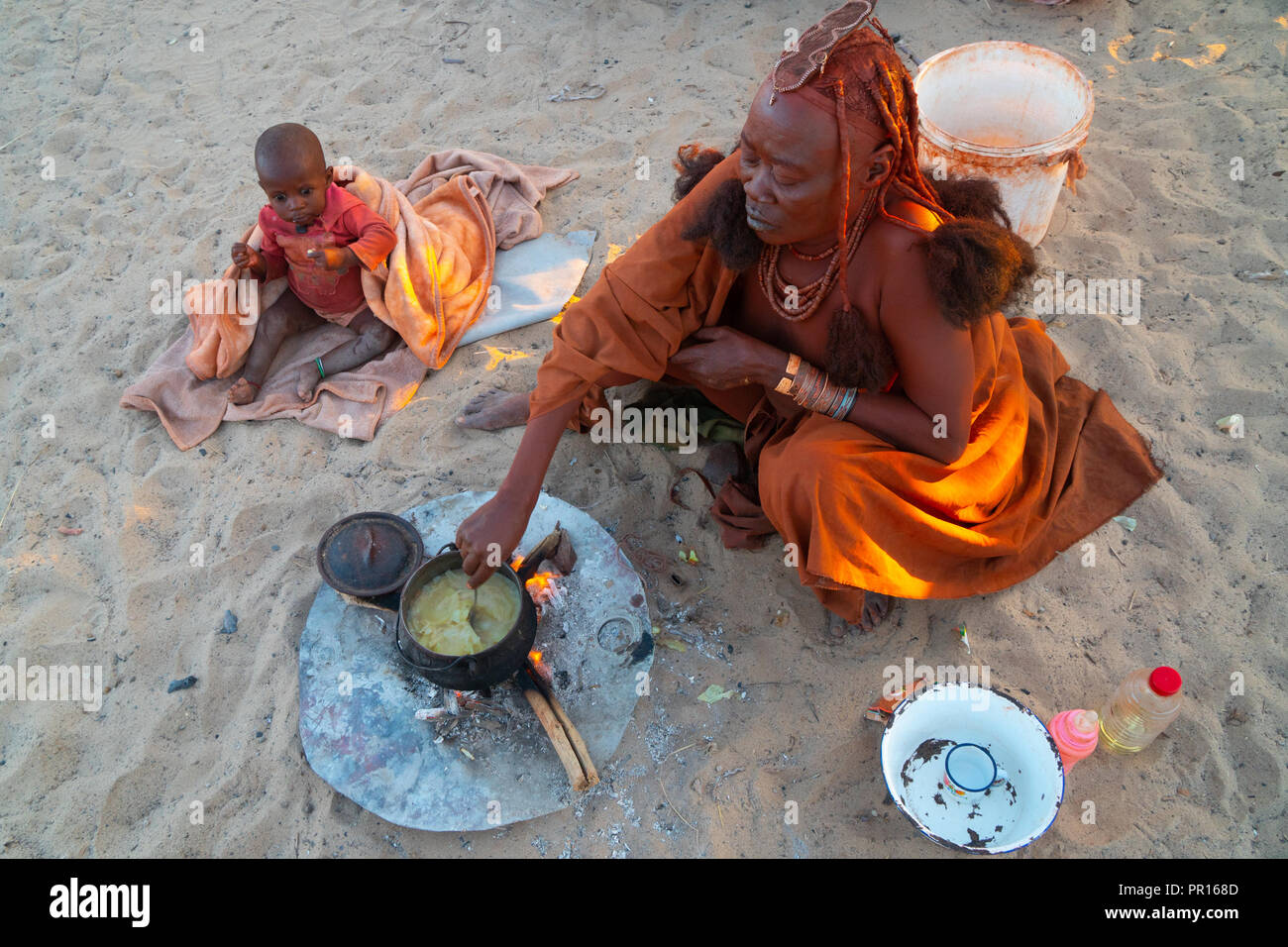 L'un rouge senior ochred femme Himba avec son enfant la cuisson sur un feu ouvert, Village Puros, nearr Sesfontein, Namibie, Afrique Banque D'Images