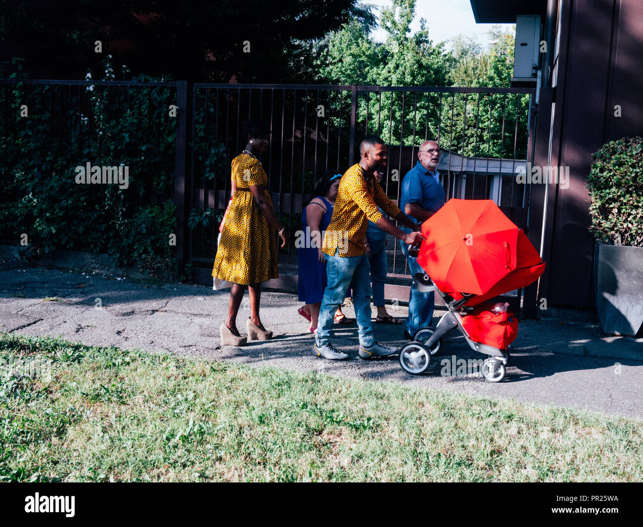 La famille noire élégante marche sur un trottoir avec une poussette dans une robe jaune vif, le tout entouré de vert. Monza, Italie. Banque D'Images